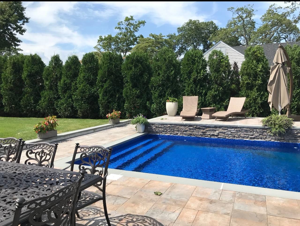 A stone pool deck with a stacked-stone veneer wall, planters, and a tall arborvitae privacy screen along the property line.