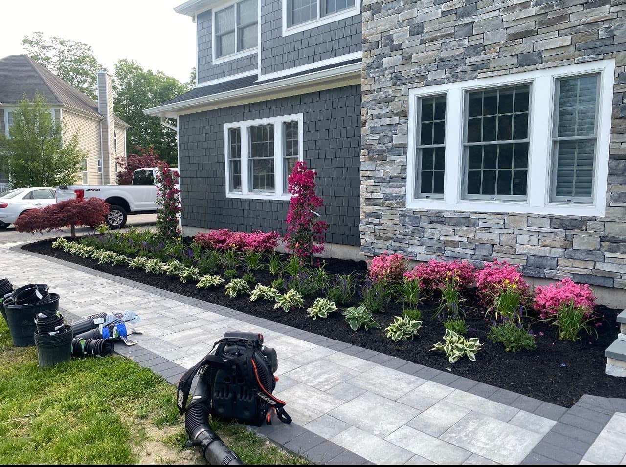 A paver walkway running alongside a stone-veneered house, with newly planted foundation beds of azaleas, hostas, and flowering vines in dark mulch.
