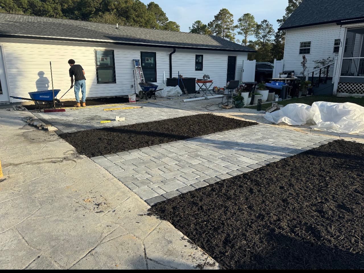 A crew member sweeping jointing sand into a newly laid paver walkway between two buildings, with fresh mulch beds on both sides.